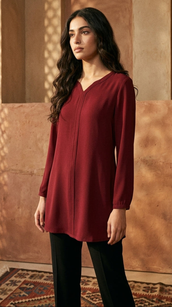 A woman modeling a long-sleeve, V-neck tunic top in deep burgundy, paired with black pants, standing in a warm, sunlit room with textured brown walls and a patterned rug.
