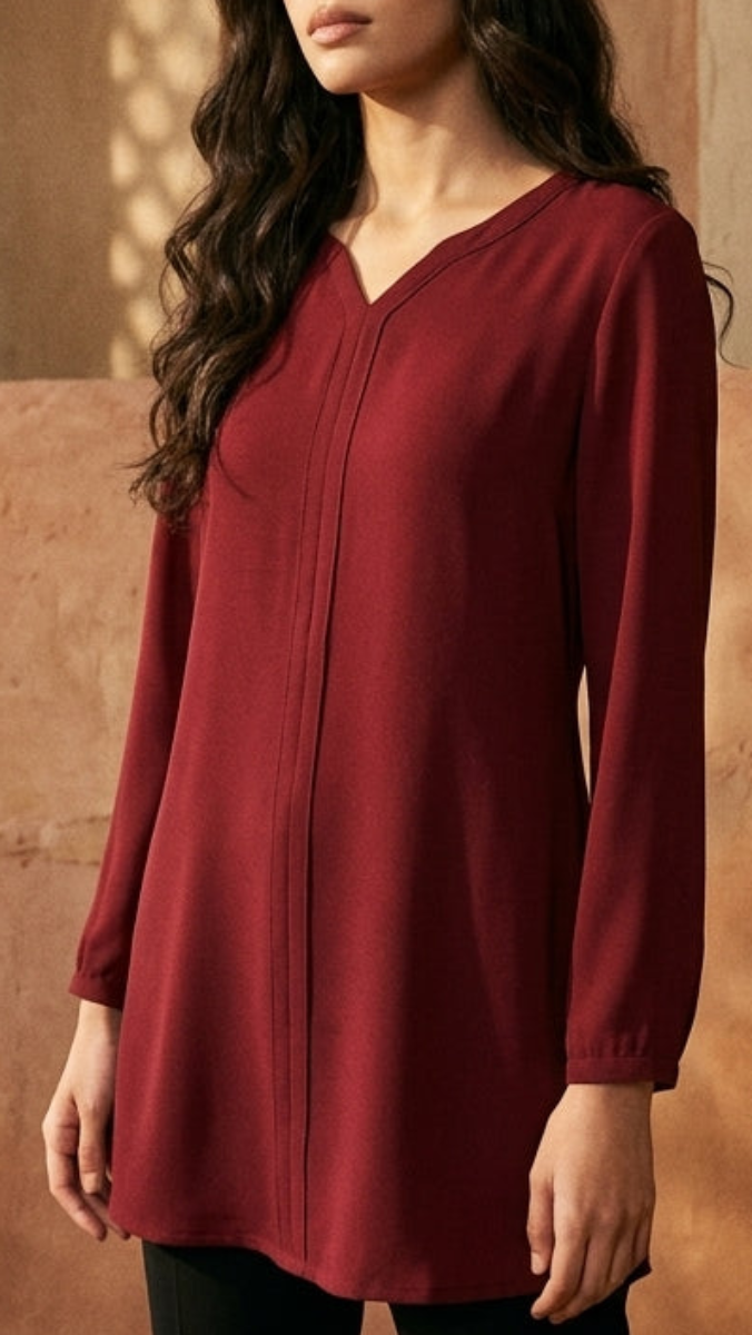 A woman modeling a long-sleeve, V-neck tunic top in deep burgundy, paired with black pants, standing in a warm, sunlit room with textured brown walls and a patterned rug.