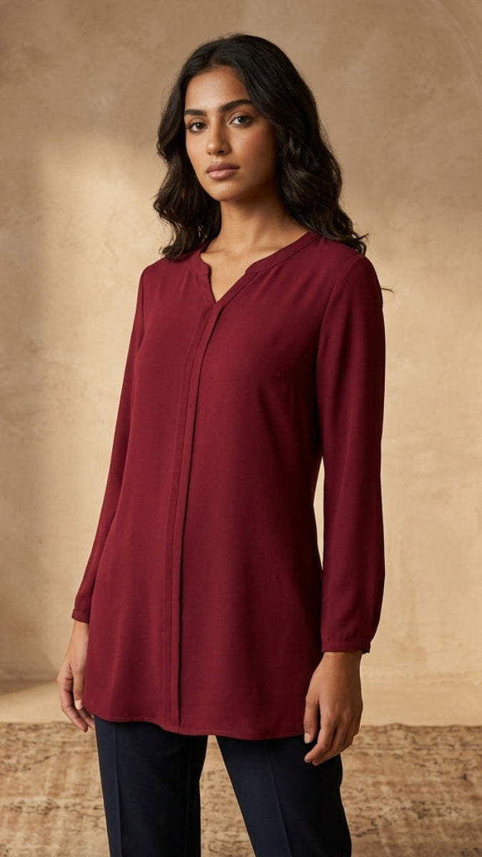 A woman modeling a long-sleeve, V-neck tunic top in deep burgundy, paired with dark pants, standing in a rustic room with arched walls, pottery, and a rug.