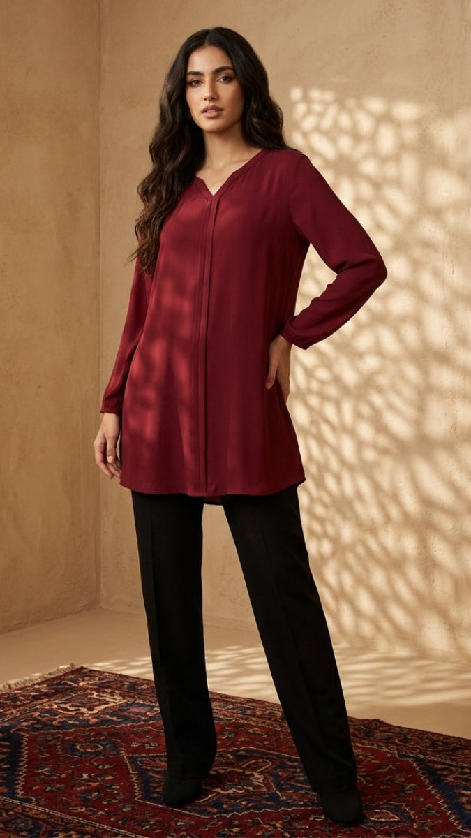 A woman modeling a long-sleeve, V-neck tunic in deep burgundy with black trousers, standing on a patterned rug against a beige wall with decorative panels and leaf shadows.