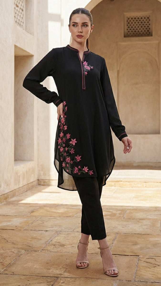 Woman in a black outfit with pink floral embroidery standing in a sunlit stone corridor.