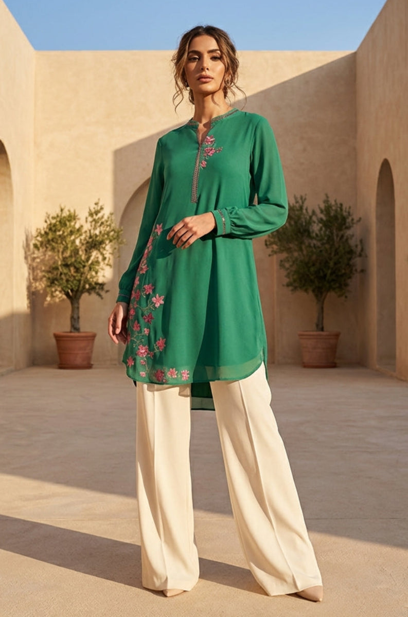 Woman in a green embroidered top and white pants standing in a sunlit courtyard with arches and potted plants.