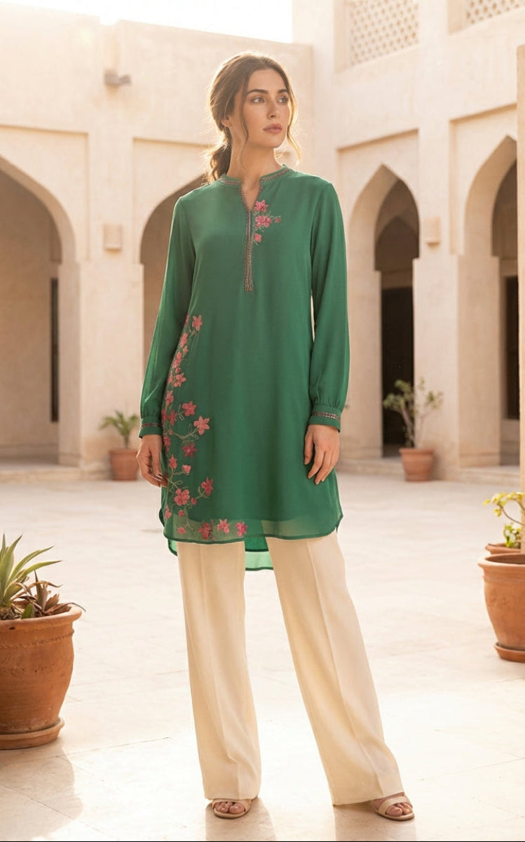 Woman in a green embroidered top and beige pants standing in a sunlit courtyard with arches and potted plants.