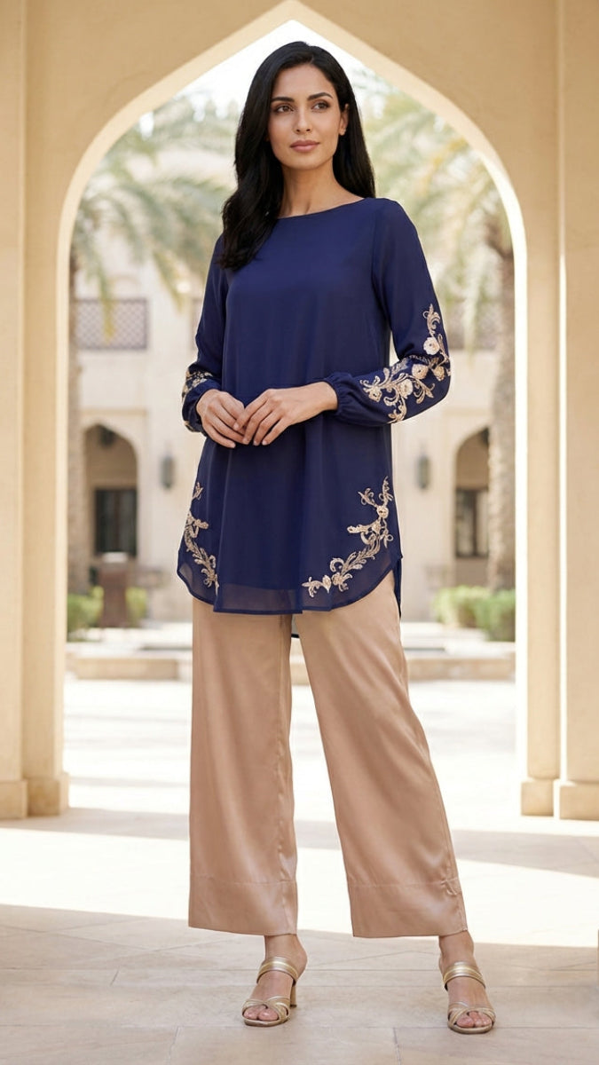 Woman wearing a blue top with floral embroidery and beige pants standing in front of an archway.