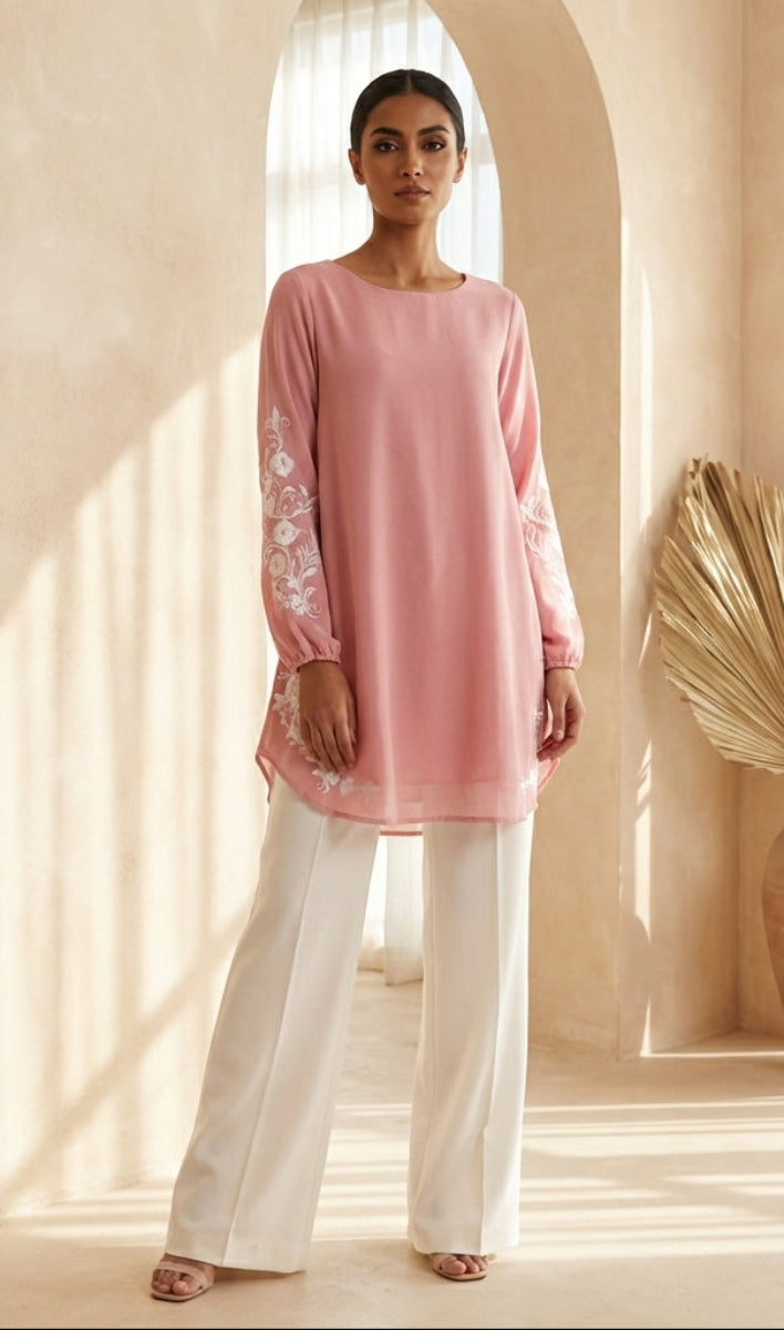 Woman wearing a pink top with lace details and white pants in a bright room.