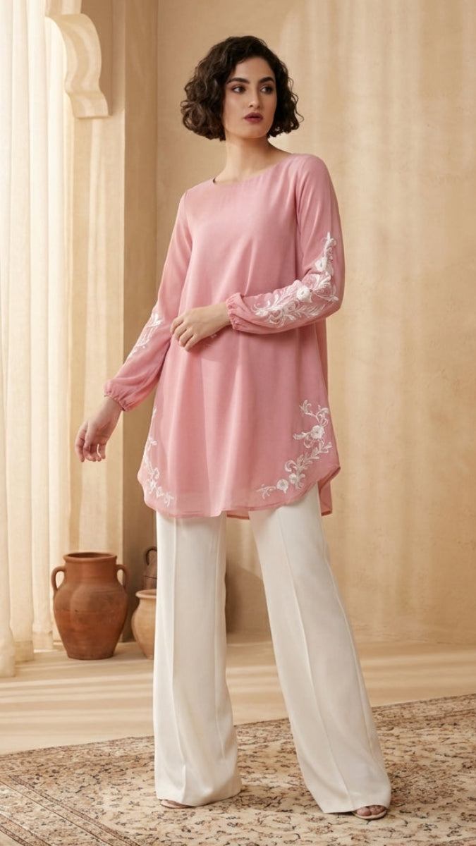 Woman wearing a pink top with lace sleeves and white pants in a room with decorative elements.