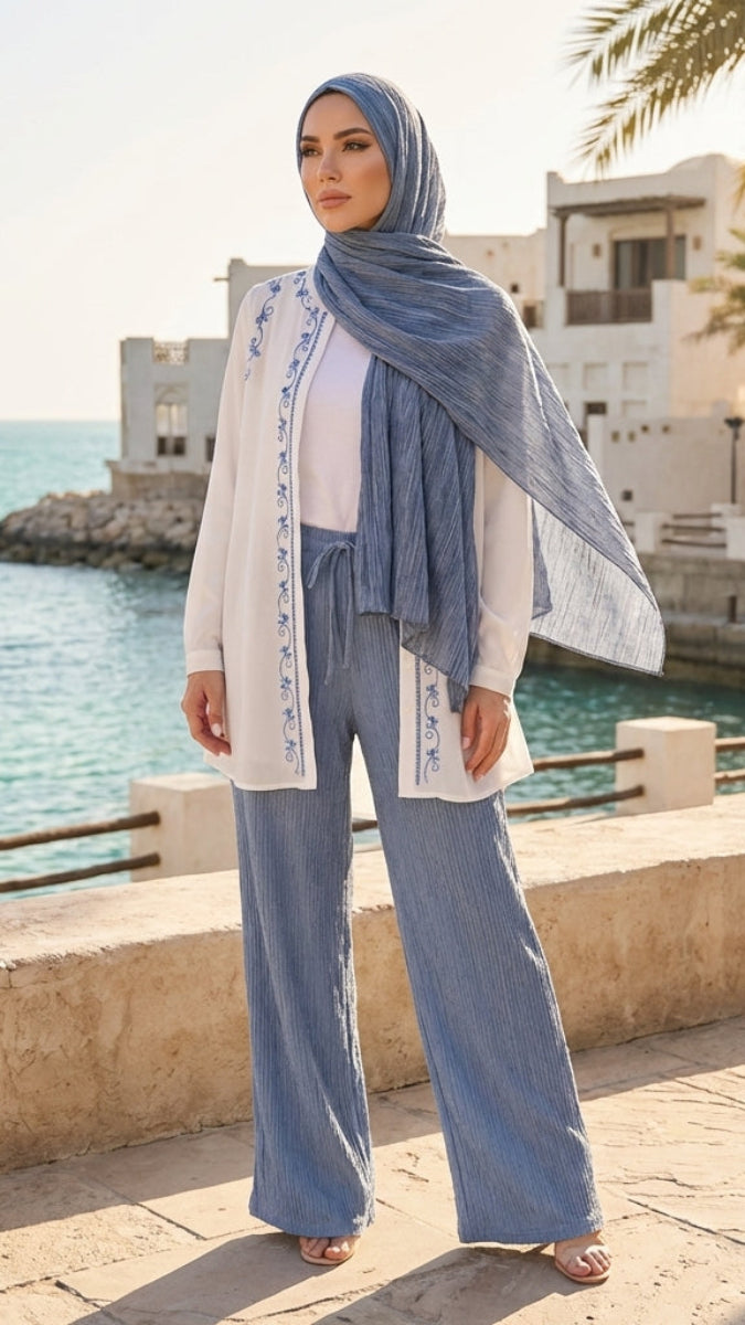 Woman in a hijab and white outfit standing by a waterfront with palm trees and buildings in the background.