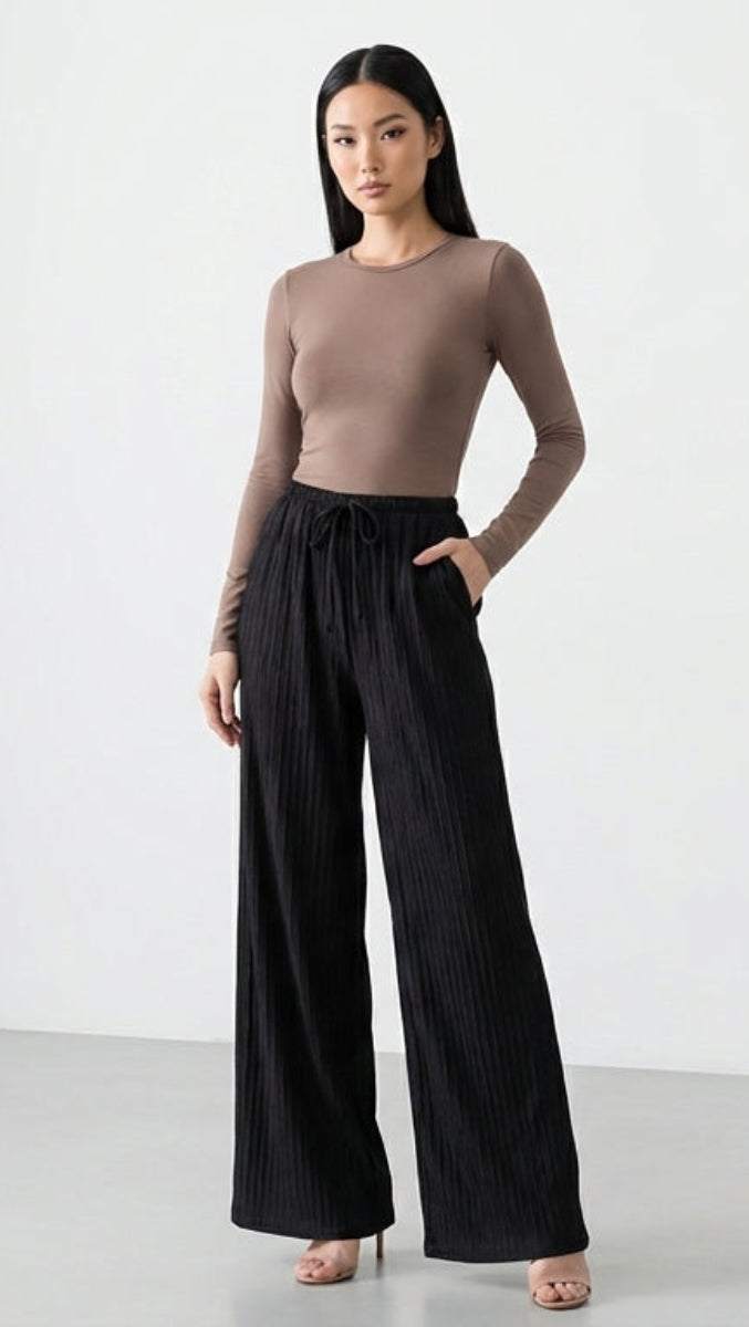 Woman wearing a brown top and black wide-leg pants in a minimal white room.