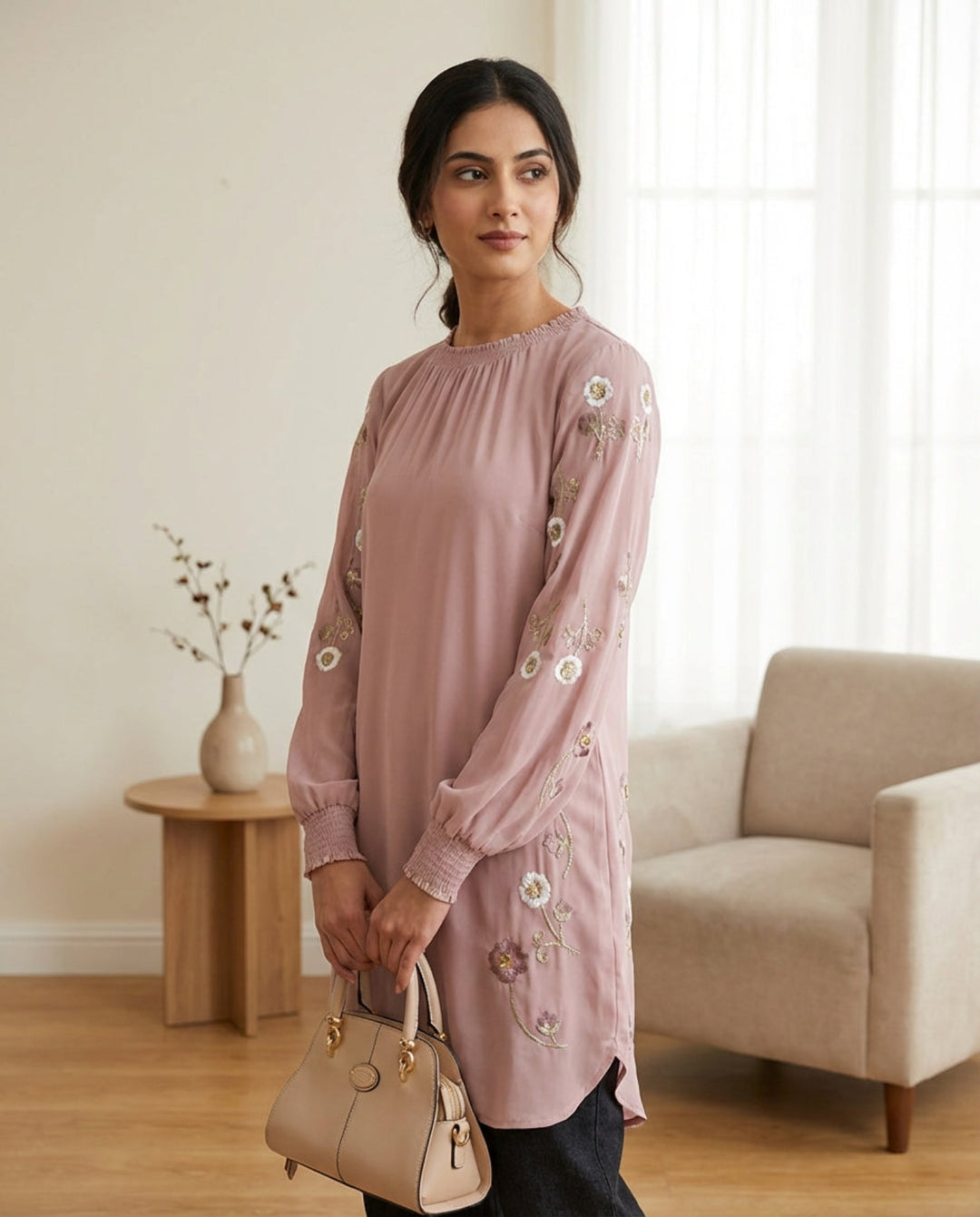 Woman in a pink embroidered dress holding a beige handbag in a living room.