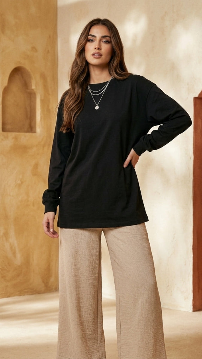 Woman wearing a black top and beige pants standing in a sunlit room with arched doorways.