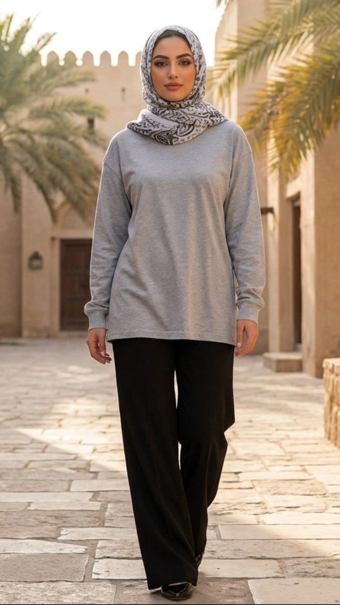 Woman in a gray top and black pants walking in a sunlit courtyard with palm trees and beige buildings.