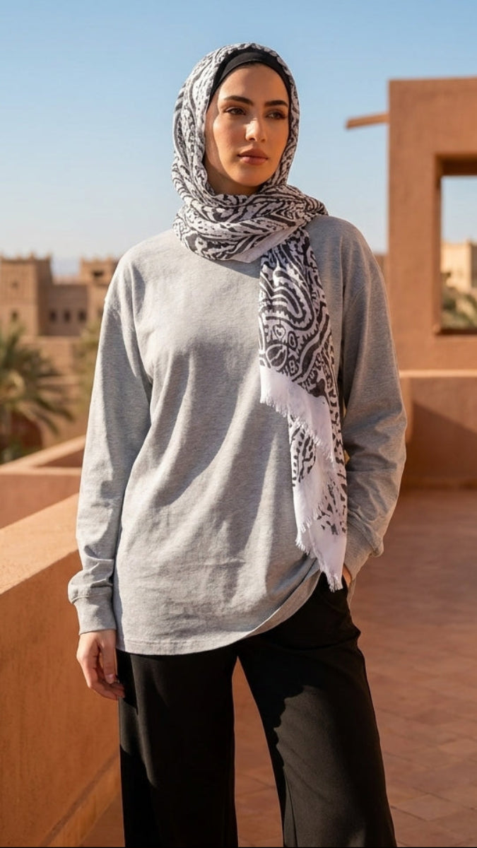 Woman wearing a patterned hijab and gray sweater standing on a sunlit terrace with desert landscape.