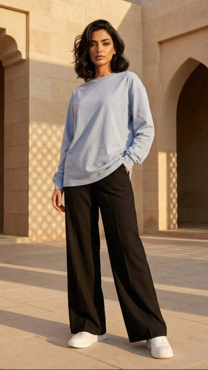 Woman standing in a sunlit architectural courtyard wearing a light blue sweater and black pants.
