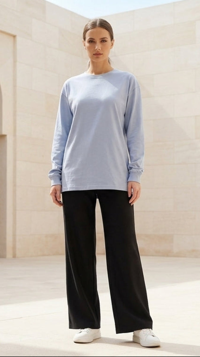 Woman standing in a sunlit architectural courtyard wearing a light blue top and black pants.