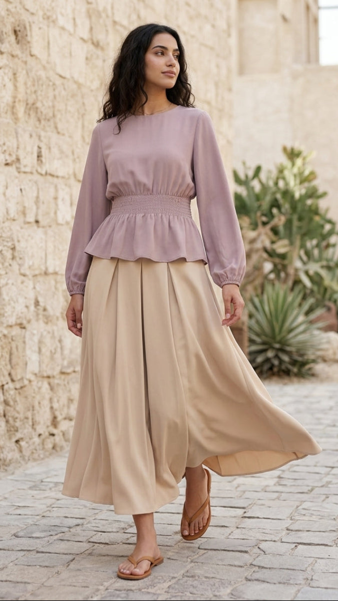 Woman wearing a light purple top and beige skirt standing in front of a stone wall.