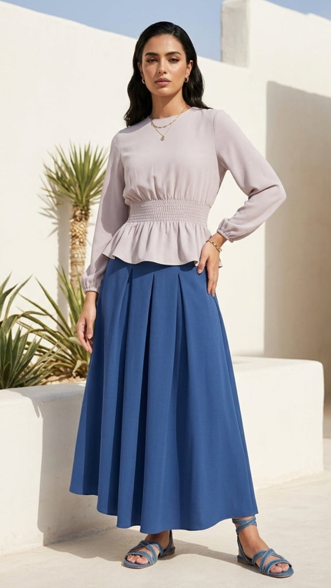 Woman in a light purple top and blue skirt standing in a sunlit outdoor setting with white architecture and greenery.