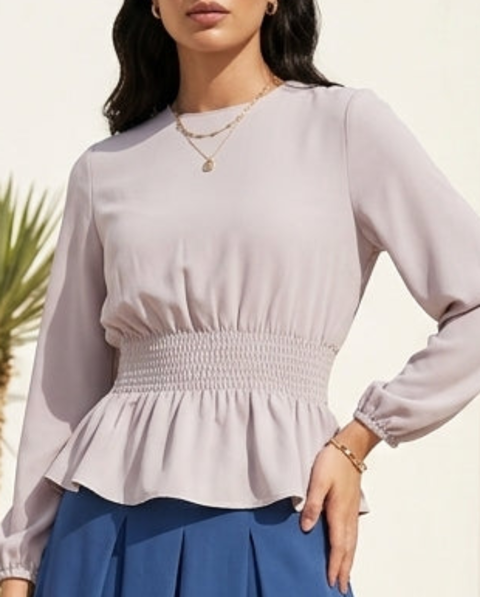 Woman in a light purple top and blue skirt standing in a sunlit outdoor setting with white architecture and greenery.
