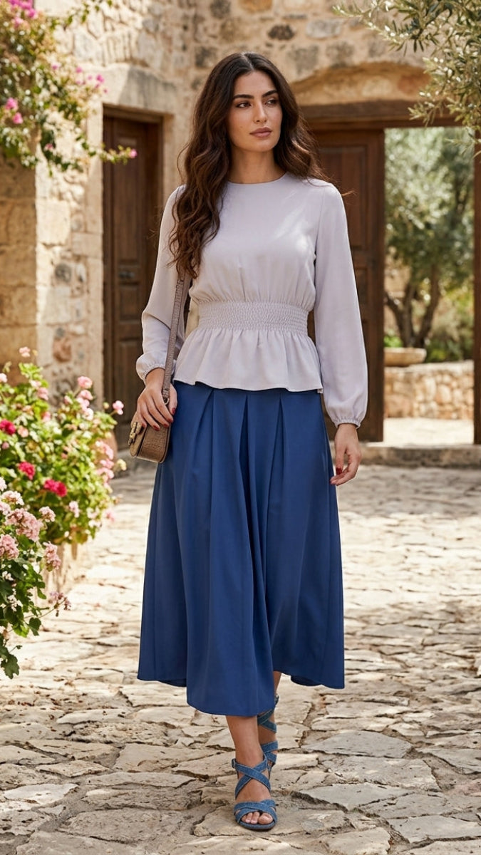 Woman in a white top and blue skirt standing on a stone path with flowers and stone buildings in the background.