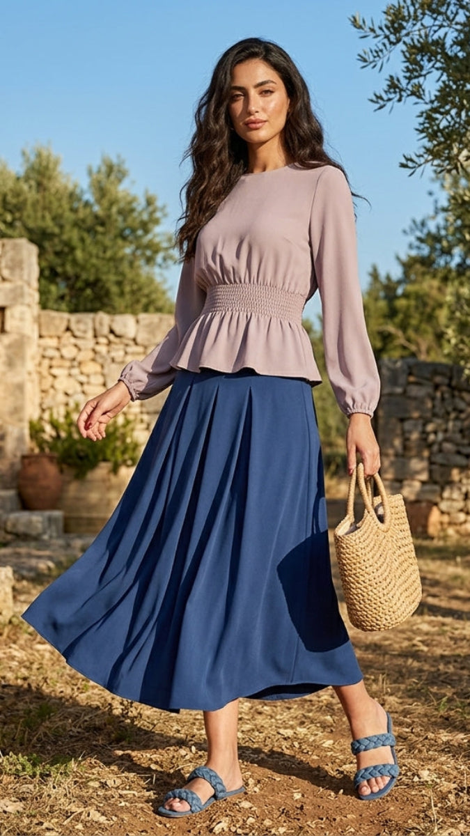 Woman in a pink top and blue skirt standing in an outdoor setting with stone walls and trees.