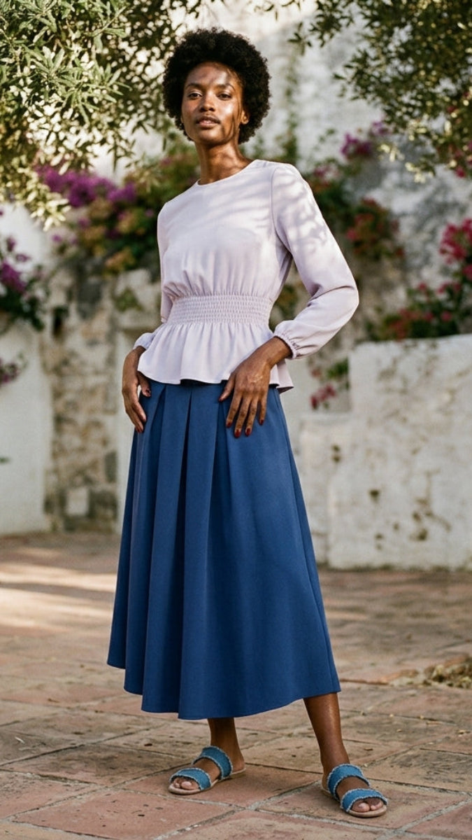 Woman in a white top and blue skirt standing in a garden with flowers and a stone wall in the background.