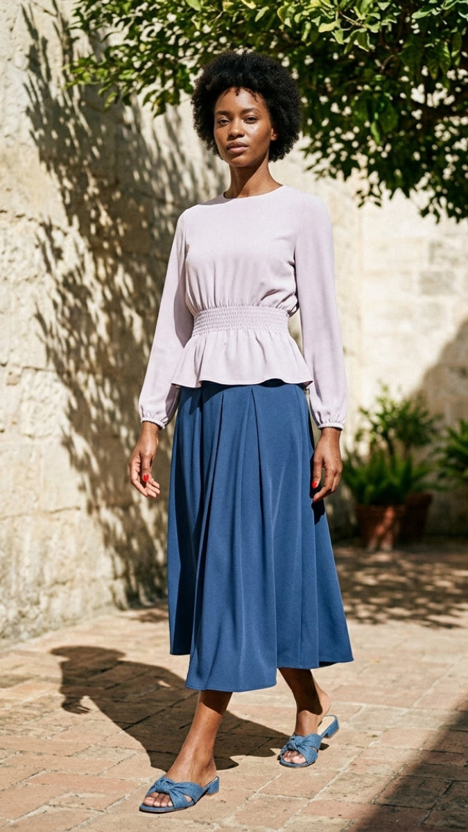 Woman wearing a light purple peplum top and blue skirt standing outdoors against a stone wall.