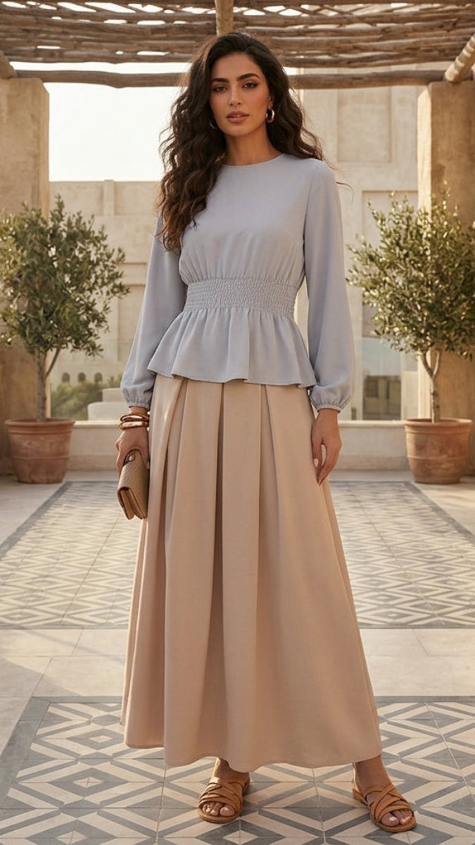 Woman in a light blue top and beige pants standing in a sunlit outdoor setting with plants and stone architecture.