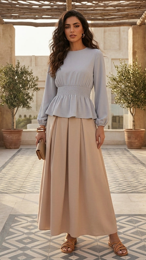 Woman in a light blue top and beige pants standing in a sunlit outdoor setting with plants and stone architecture.