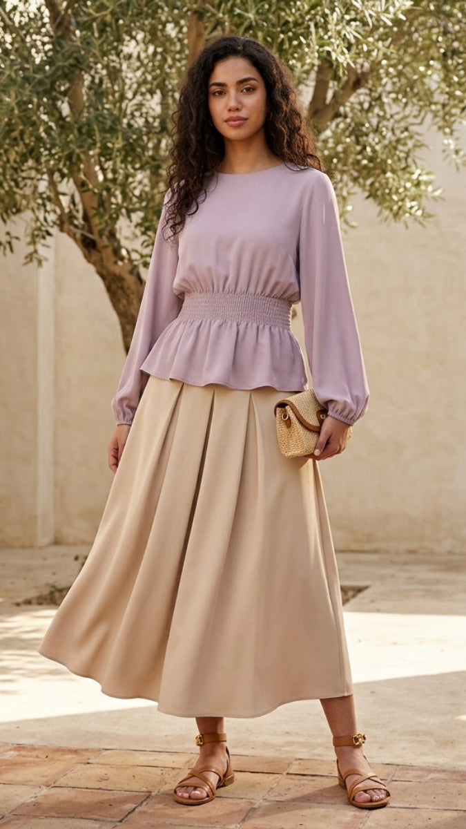 Woman in a light purple top and beige skirt standing in a sunlit outdoor setting.