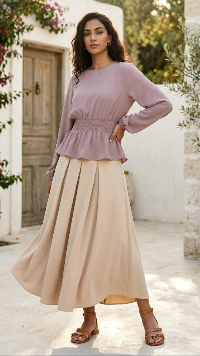 Woman wearing a mauve top and beige skirt standing in a sunlit outdoor setting.