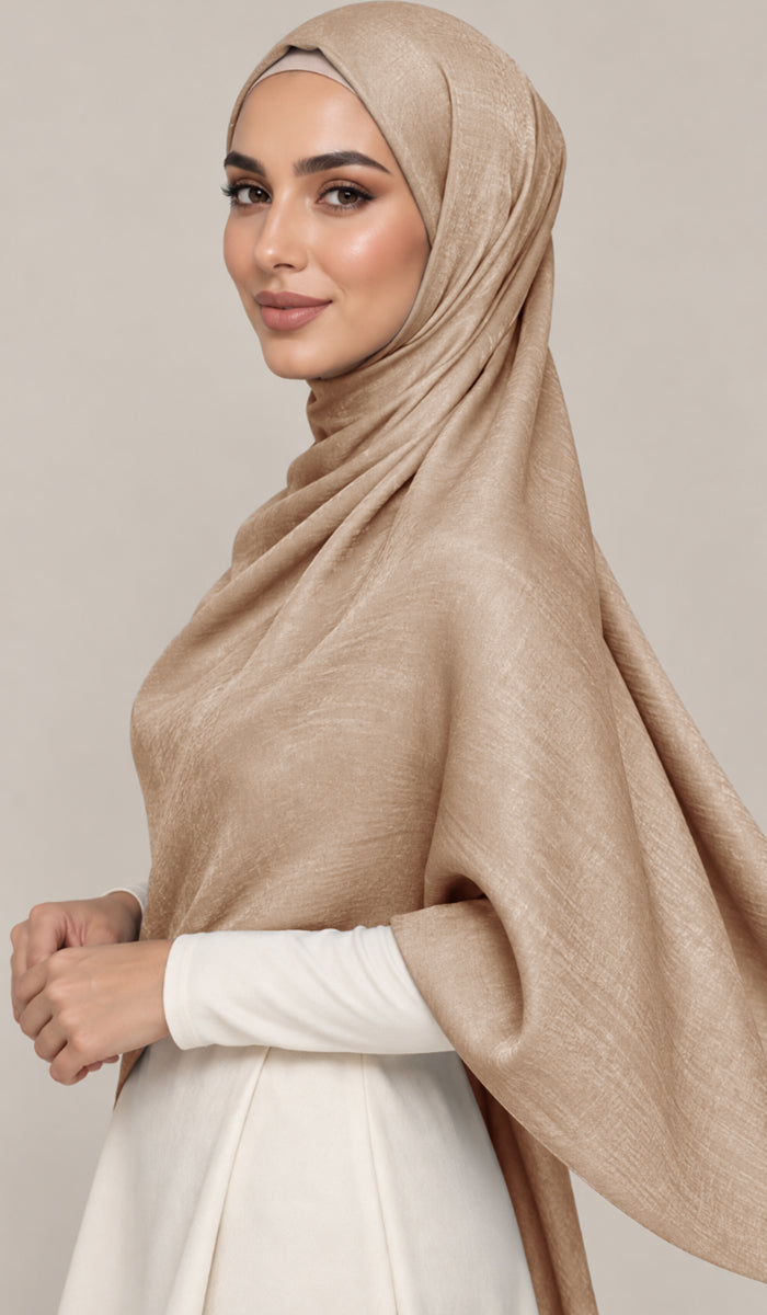 A woman wearing a draped beige shawl over her shoulders and a white long-sleeve dress, posed against a solid light gray studio background.