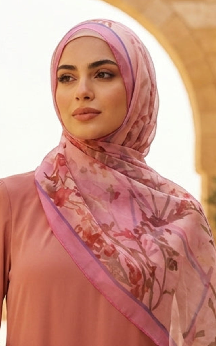 Woman in a pink dress and headscarf standing in a sunlit courtyard with arches and potted plants.