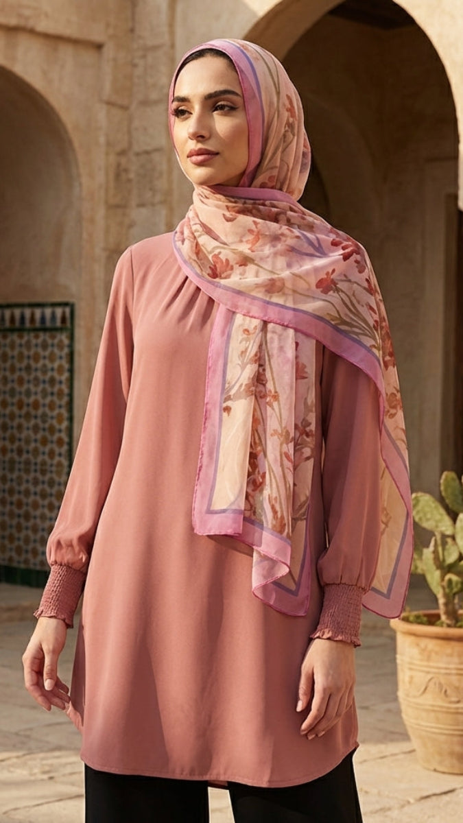 Woman wearing a pink dress and floral hijab standing in a courtyard with stone arches and potted plants.