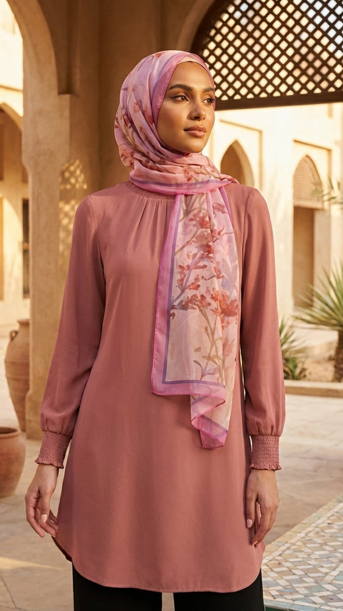 Woman wearing a pink dress and floral hijab in an outdoor setting with arches and plants.