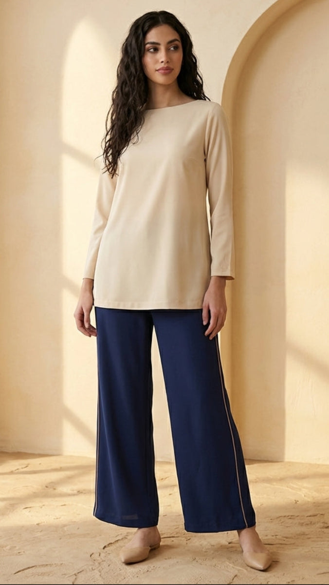 Woman wearing a beige top and blue pants standing in a sunlit room with a vase of dried plants.
