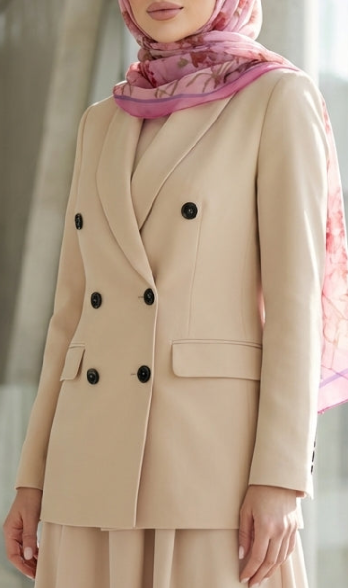 Woman in a beige suit and pink hijab standing in a modern indoor setting.