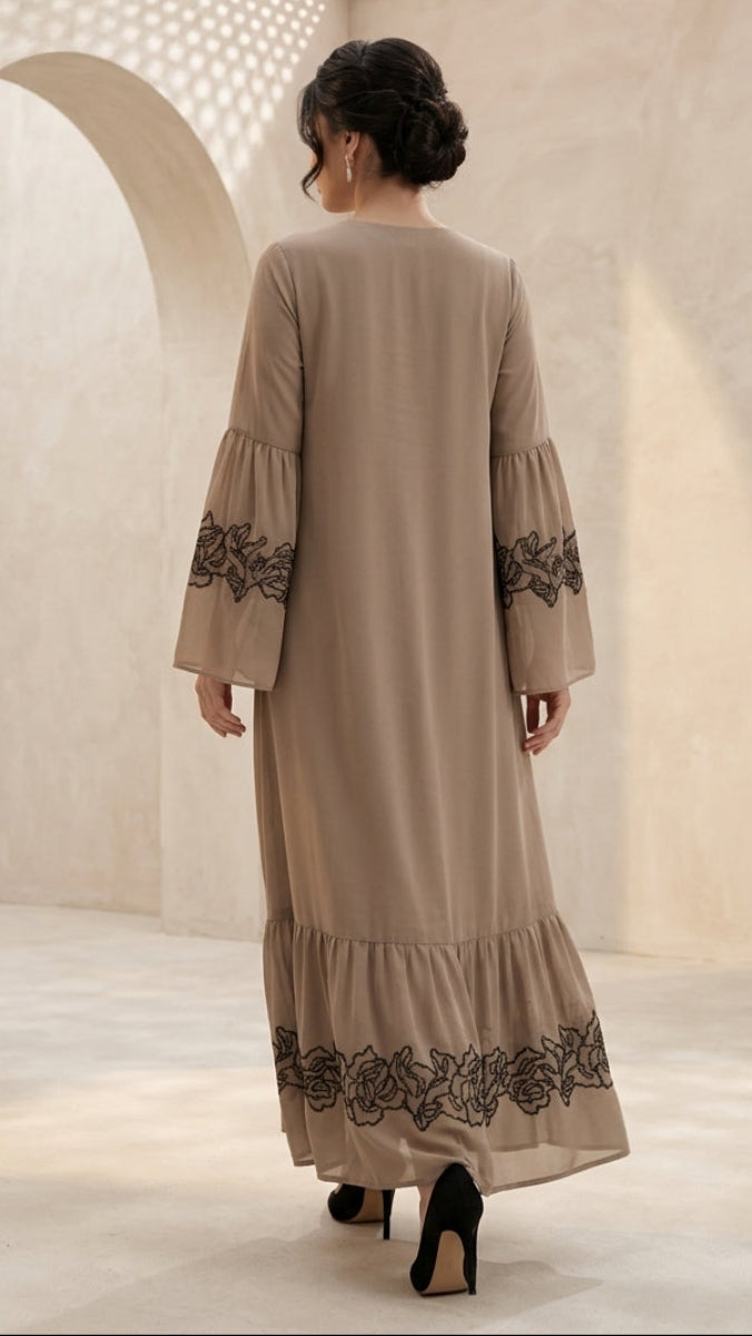 A woman viewed from behind wearing a beige long-sleeved maxi dress with black floral lace embroidery on the cuffs and hem, standing in a warm-toned architectural setting with arches.