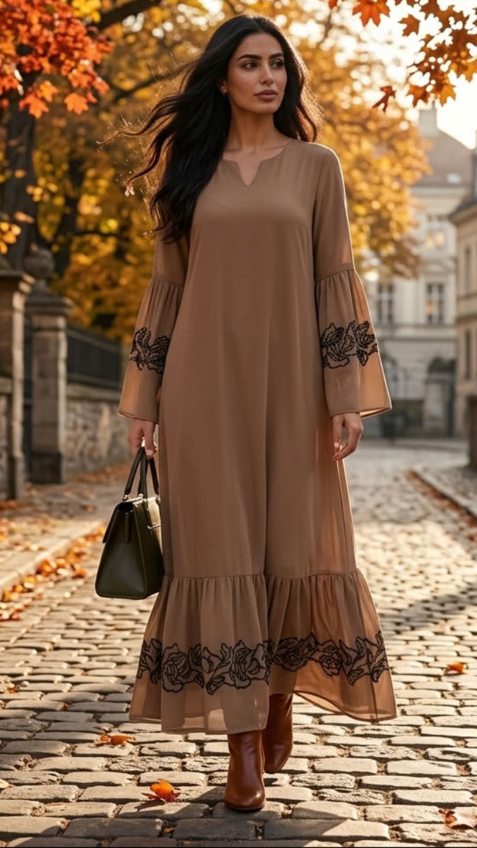 A woman wearing a long taupe-brown maxi dress with black floral lace trim on the sleeves and hem, walking down a cobblestone street lined with autumn trees and fallen leaves.