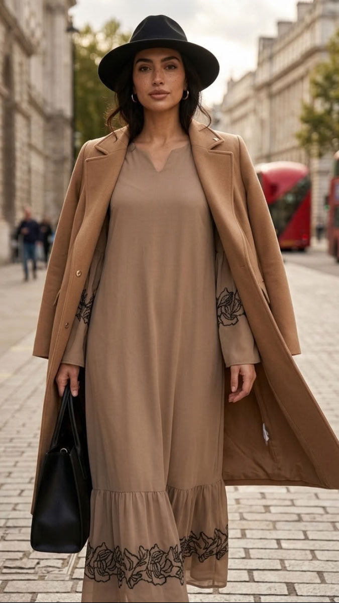 A woman stands on a city street wearing a camel trench coat over a matching beige maxi dress with black floral embroidery at the hem and cuffs, along with a black wide-brim hat and a black handbag.