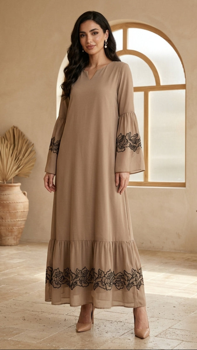 A woman models a long beige maxi dress with black floral embroidery on the sleeves and hem, standing in a warm, neutral-toned room with a large arched window.