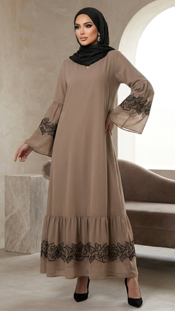 A woman wearing a long taupe-beige maxi abaya with black floral lace embroidery on the sleeves and hem, paired with a black hijab and black heels, standing in a modern minimalist interior with a curved sofa.