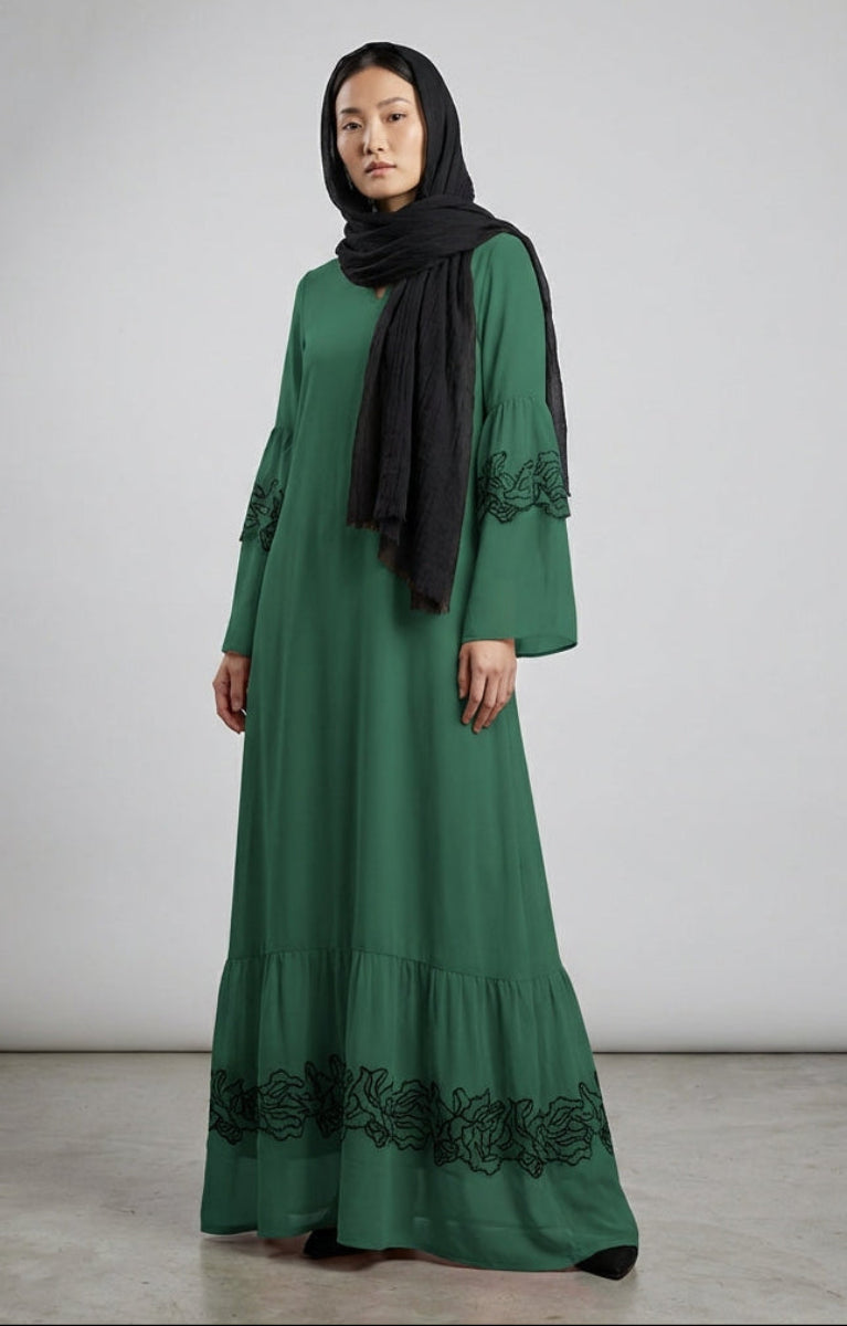 Woman wearing a green dress with black embroidery and a black hijab against a white background