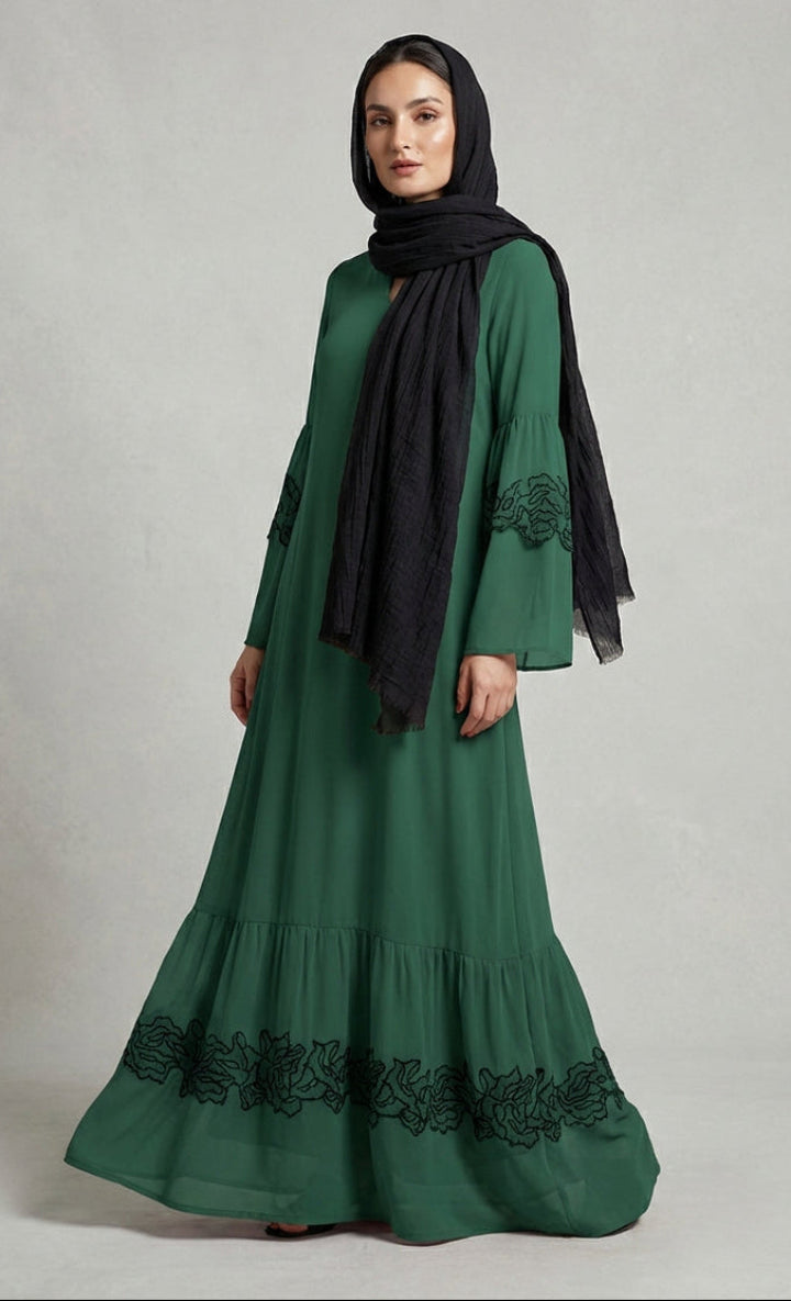 Woman wearing a green dress with black embroidery and a black hijab on a gray background