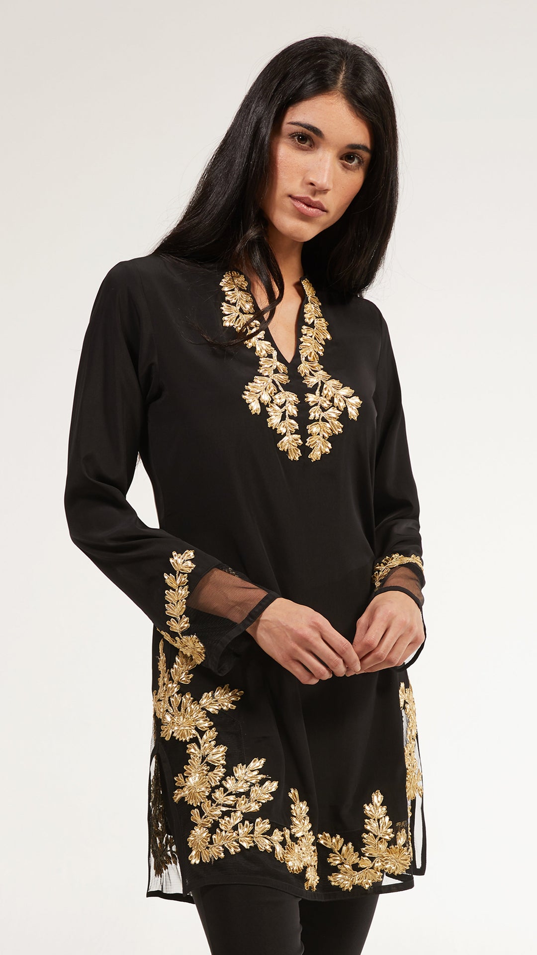 A woman modeling a black long-sleeve tunic with ornate gold floral embroidery around the neckline, cuffs, and hem, photographed against a plain light gray studio background.