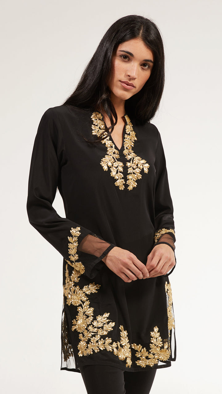 A woman modeling a black long-sleeve tunic with ornate gold floral embroidery around the neckline, cuffs, and hem, photographed against a plain light gray studio background.