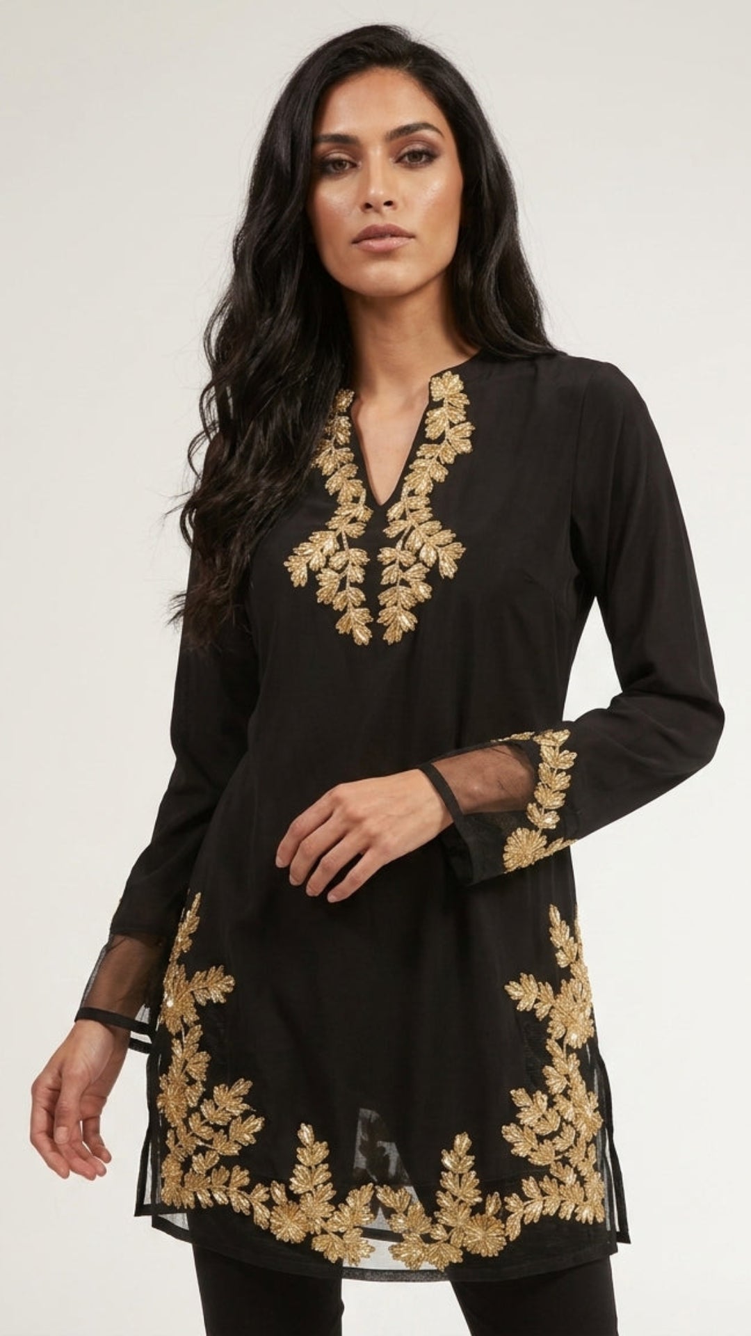 A woman modeling a black long-sleeve tunic with ornate gold floral embroidery around the neckline, cuffs, and hem, photographed against a plain light gray studio background.