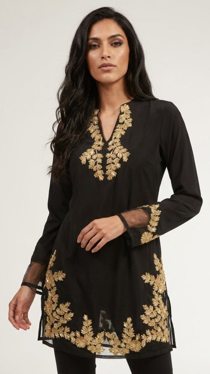 A woman modeling a black long-sleeve tunic with ornate gold floral embroidery around the neckline, cuffs, and hem, photographed against a plain light gray studio background.