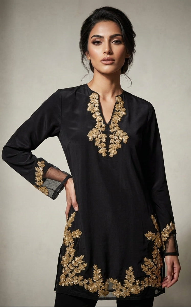 A woman modeling a black long-sleeve kurta with ornate gold floral embroidery around the V-neckline and hem, posed against a textured neutral studio backdrop.