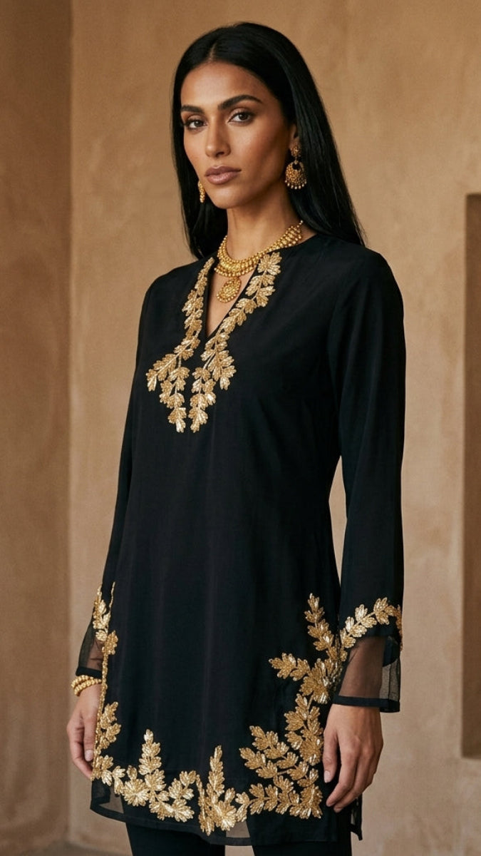 A woman modeling a black long-sleeve kurta with ornate gold floral embroidery around the neckline, cuffs, and hem, set against a warm beige architectural background.