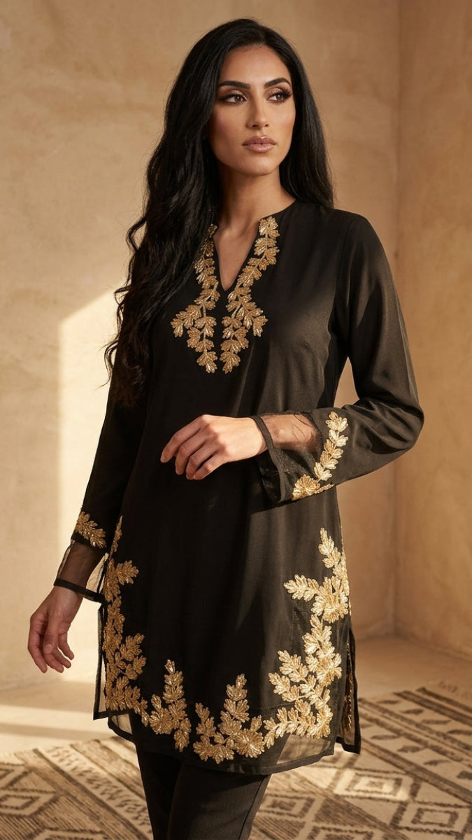 A woman modeling a black long-sleeve kurta with ornate gold floral embroidery, set against a warm, textured beige wall with soft window light.