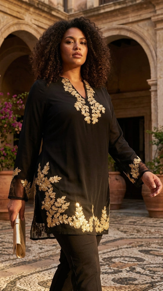 A woman stands in a stone courtyard wearing a black tunic with ornate gold floral embroidery around the neckline, hem, and cuffs. She is holding a small metallic clutch.